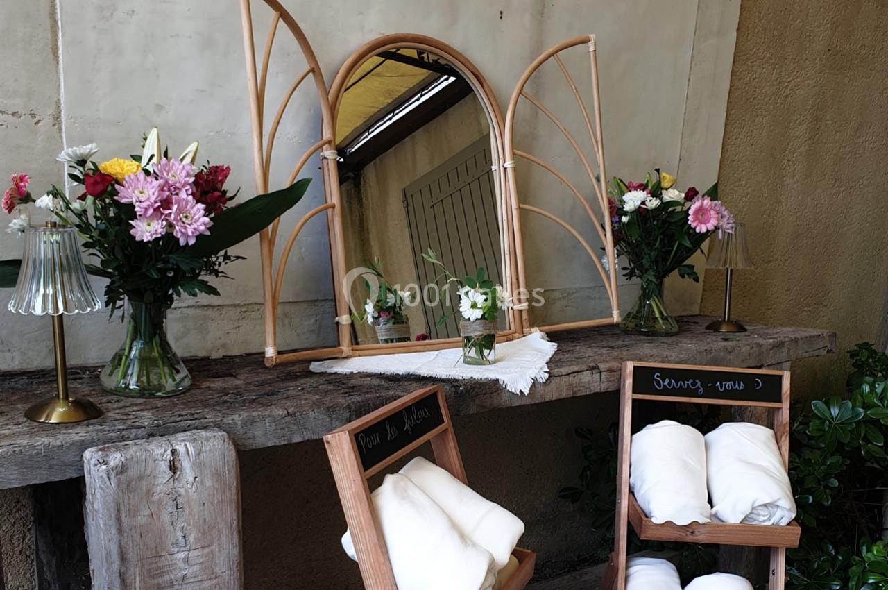 Miroir en rotin sur une table en bois, entouré de fleurs dans des vases et de chaises avec des serviettes pliées.
