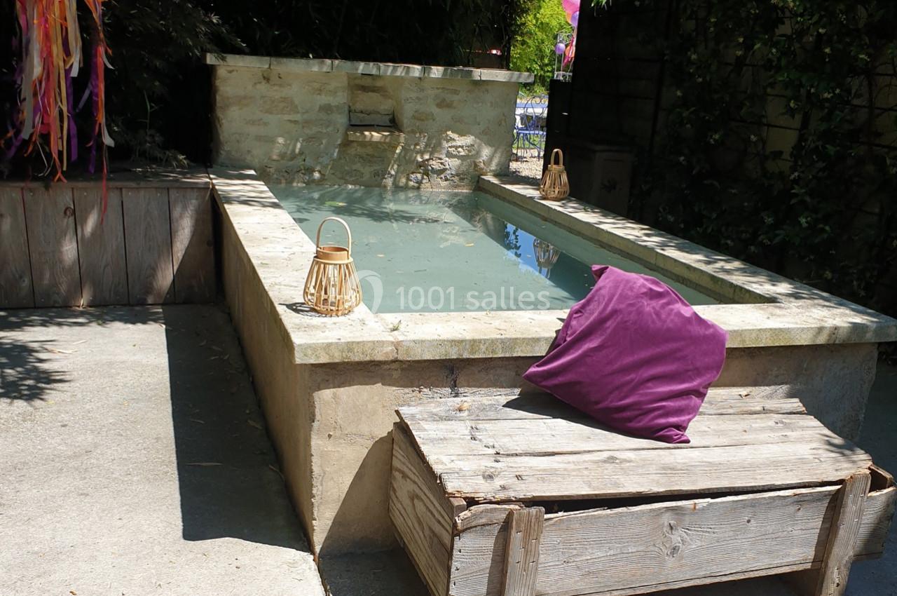 Petit bassin rectangulaire en pierre entouré de végétation, avec un banc en bois et un coussin violet au premier plan.