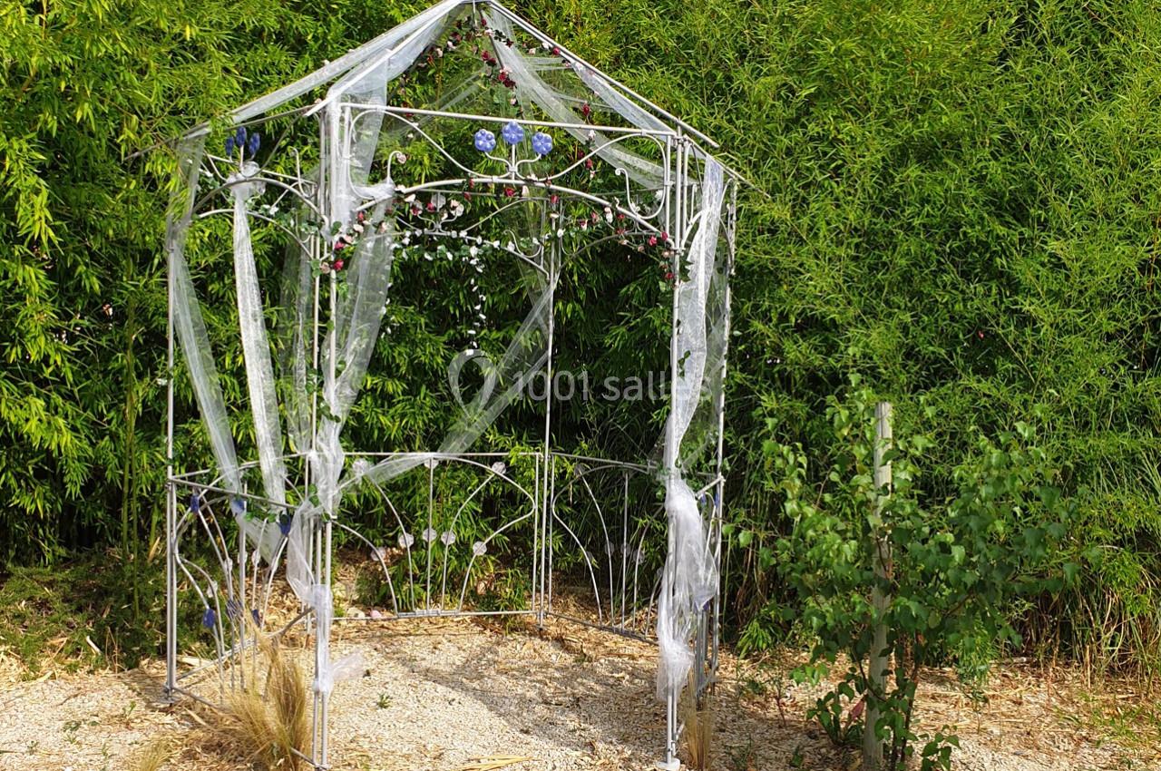 Structure métallique décorée de voiles blancs et de fleurs bleues, entourée de végétation dense.