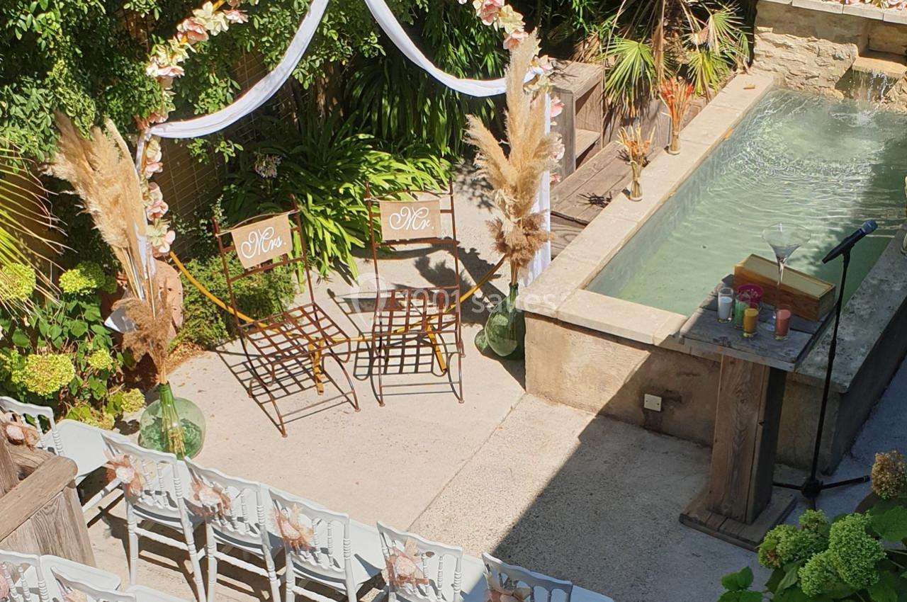 Chaises décorées sous une arche fleurie près d'un bassin, disposées pour une cérémonie en extérieur.