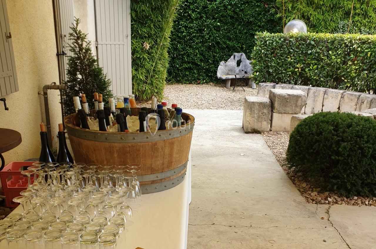 Un seau en bois rempli de bouteilles de vin avec des verres alignés sur une table, dans une cour verdoyante.