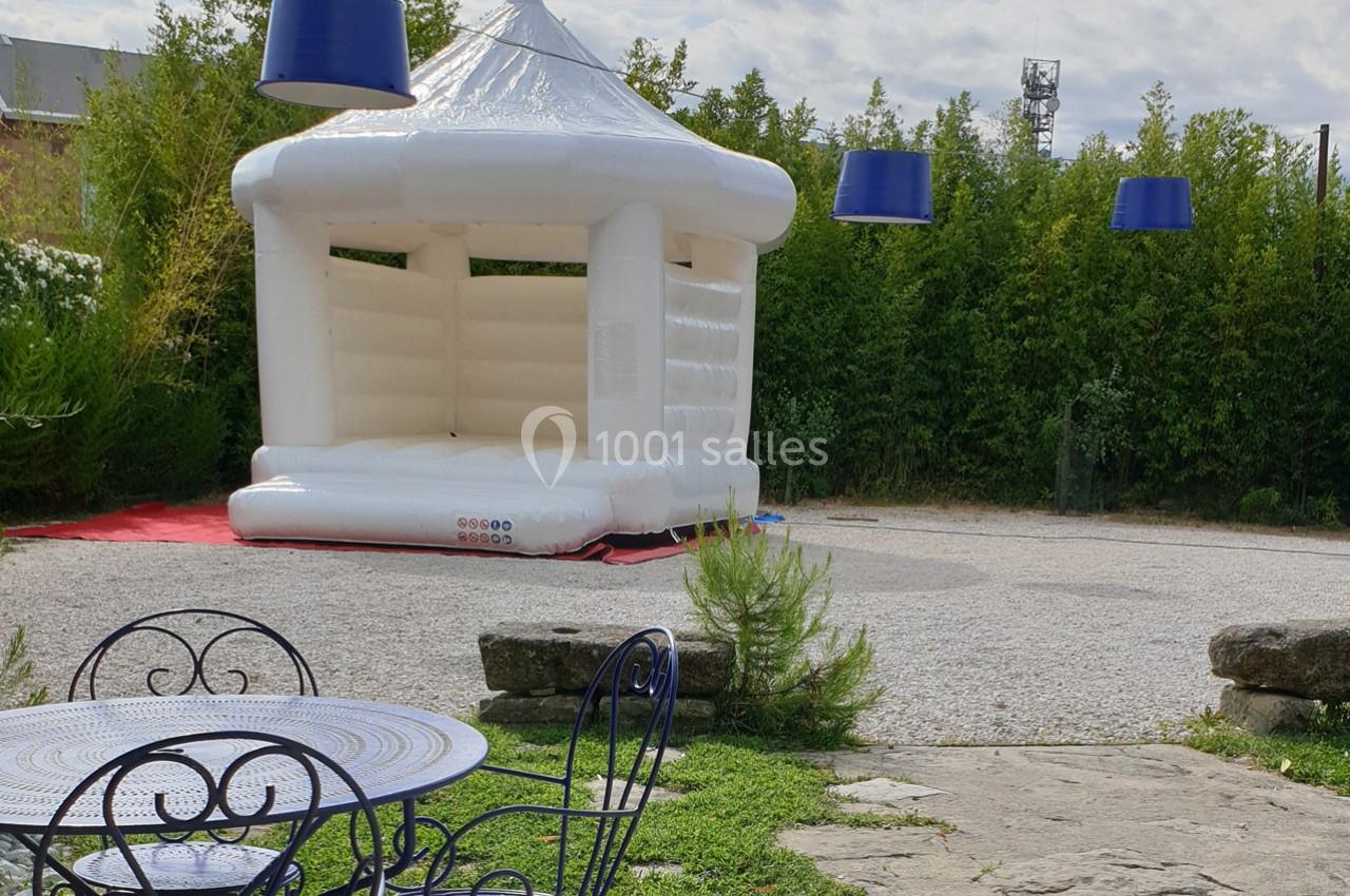 Château gonflable blanc installé sur une surface gravillonnée, entouré de végétation et de mobilier de jardin.