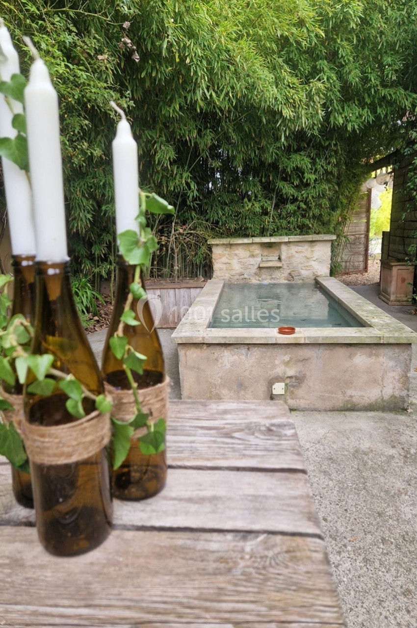 Bougies dans des bouteilles en verre décorées de lierre sur une table en bois, avec un bassin en pierre en arrière-plan.