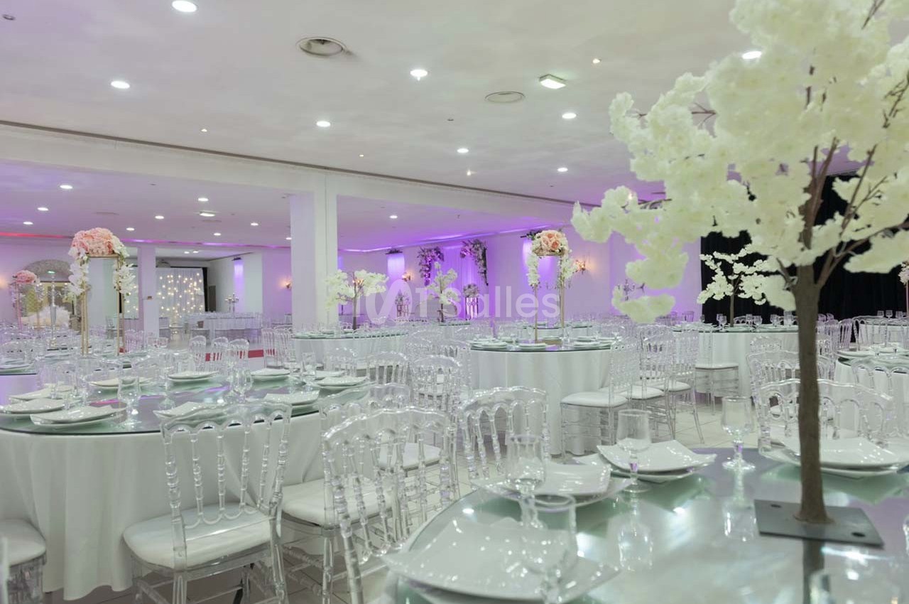 Salle de réception élégante avec tables rondes dressées, chaises transparentes et décorations florales blanches et roses.