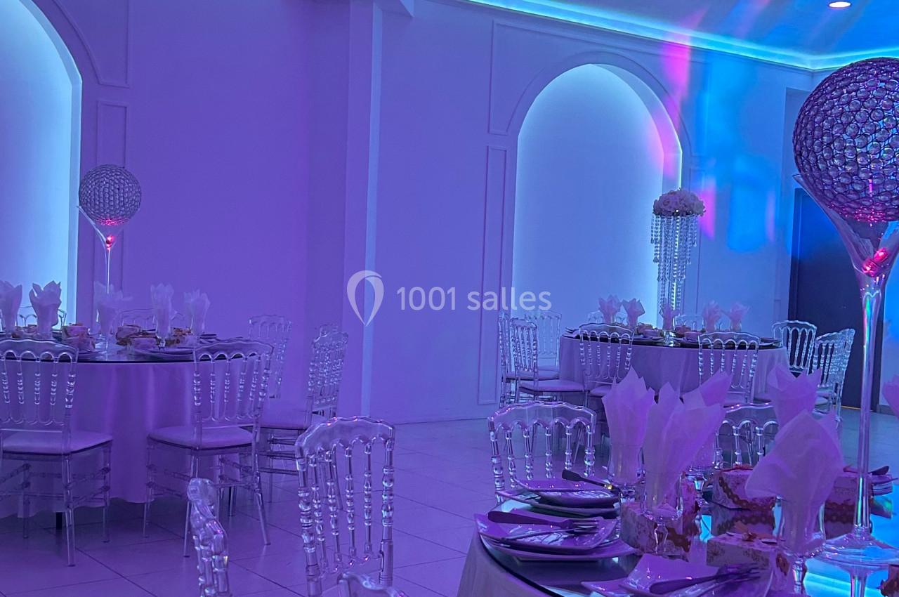 Salle de réception éclairée en violet avec tables dressées, chaises transparentes et décorations élégantes.