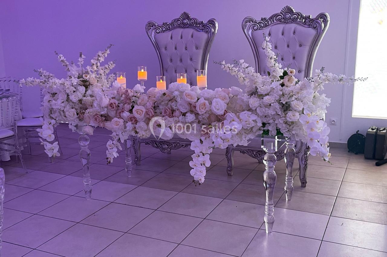 Table décorée de fleurs blanches et roses avec des bougies allumées, devant deux fauteuils argentés ornés.