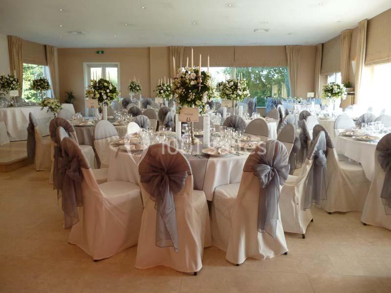 Salle de réception décorée pour un mariage avec des tables dressées, des chaises ornées de nœuds gris et des compositions…