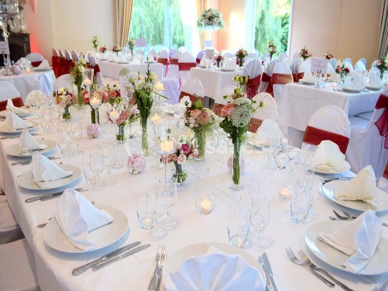 Tables élégamment dressées pour un événement, avec nappes blanches, fleurs, bougies et chaises décorées de rubans rouges.
