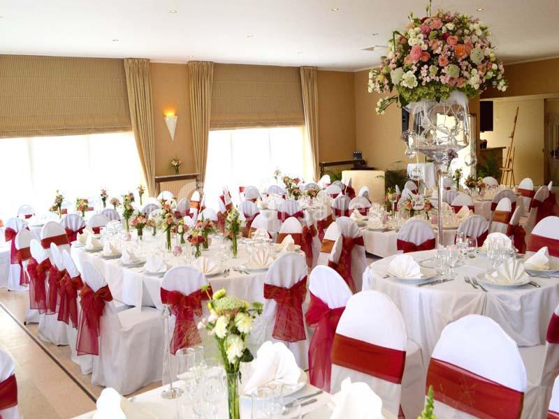 Salle de réception décorée avec des tables rondes, nappes blanches, chaises ornées de rubans rouges et centre de table…