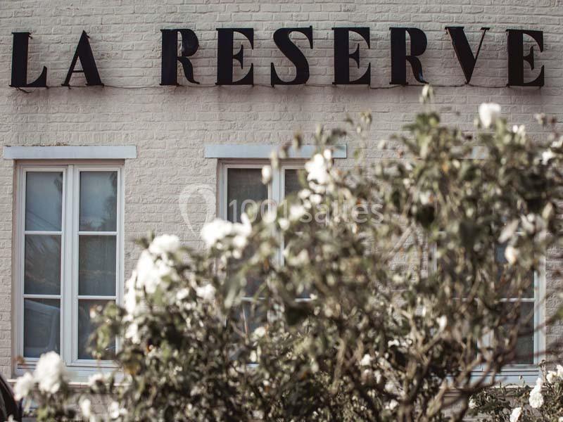 Façade d'un bâtiment en briques blanches avec des fenêtres et l'inscription ’La Réserve’, entourée de végétation.