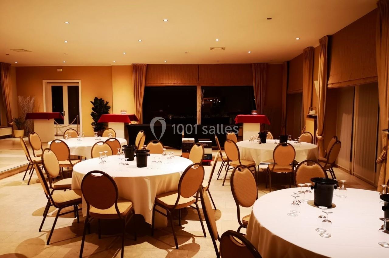 Salle de réception avec tables rondes dressées, nappes blanches, chaises en bois et grandes baies vitrées en arrière-plan.