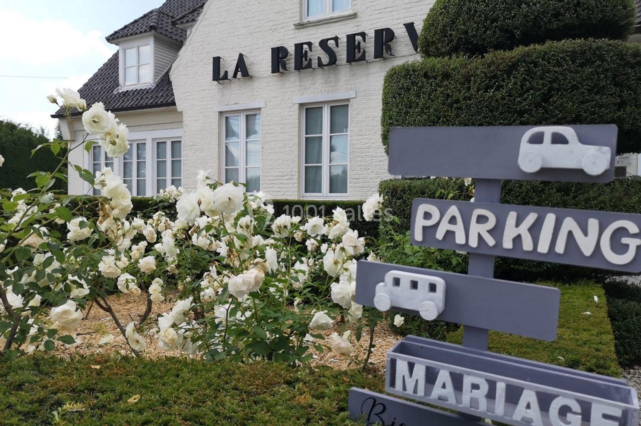Façade d'un bâtiment blanc avec des rosiers en premier plan et un panneau indiquant ’Parking Mariage’.