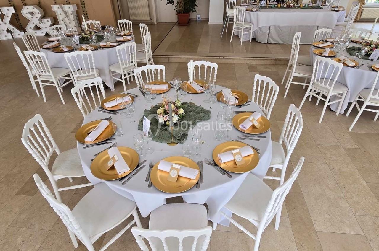 Salle de réception avec tables rondes dressées, nappes blanches, assiettes dorées et décoration florale centrale.