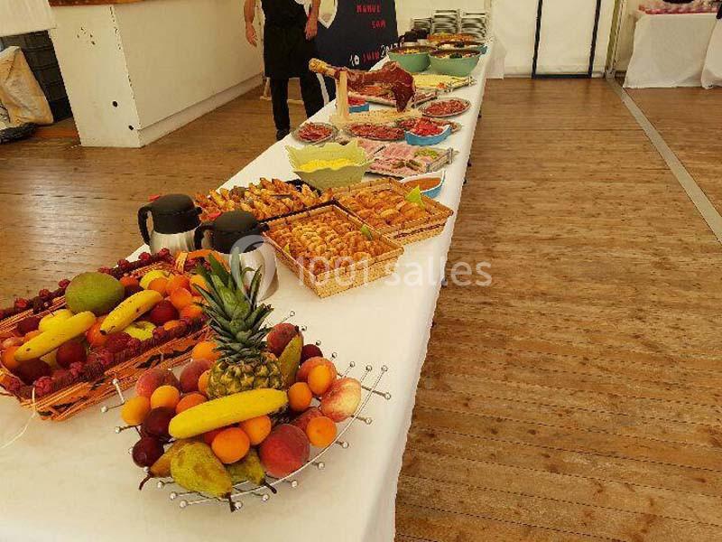 Table garnie de fruits frais, viennoiseries, charcuteries et boissons dans une salle au sol en bois.