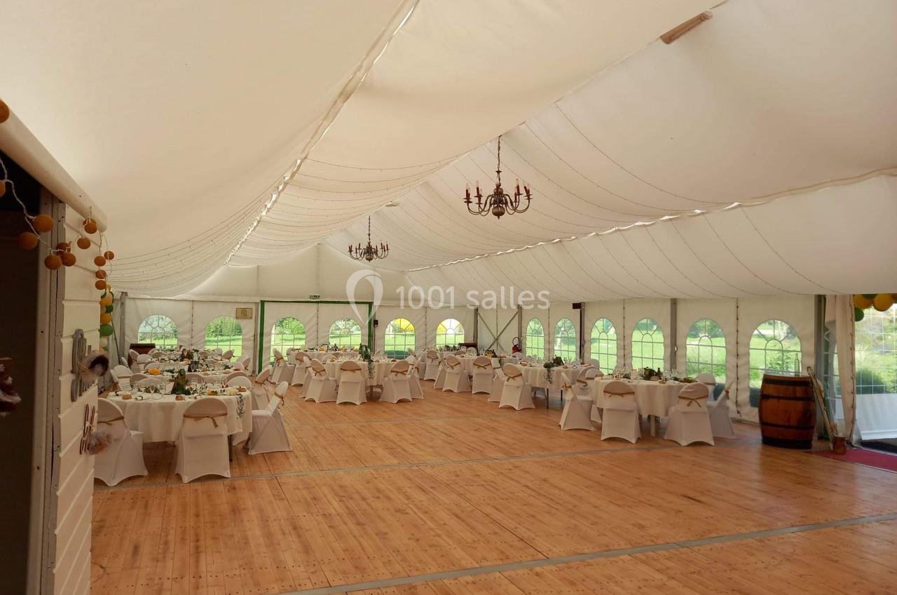 Salle de réception sous chapiteau avec tables dressées, chaises blanches et parquet en bois, éclairée par des lustres.