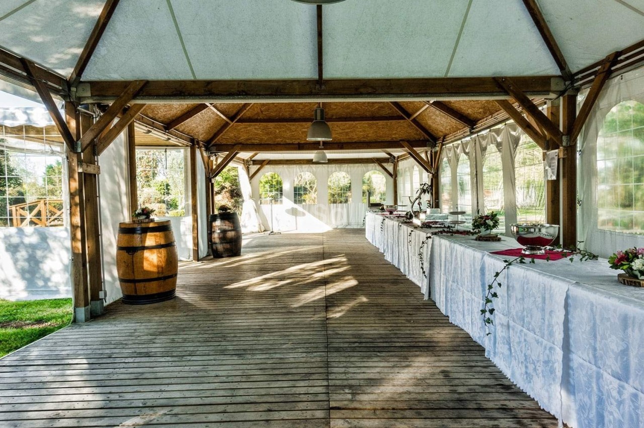 Salle de réception en bois avec tables dressées, nappes blanches et tonneaux décoratifs sous une tente ouverte.