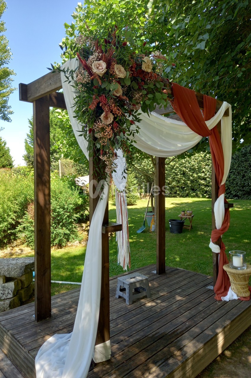 Arche en bois décorée de fleurs, tissus blancs et rouges, installée dans un jardin verdoyant.