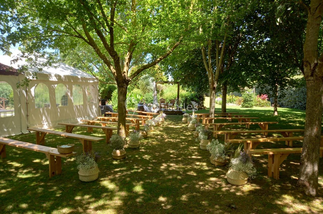 Allée centrale bordée de bancs en bois et de plantes, menant à une petite scène en plein air sous des arbres.