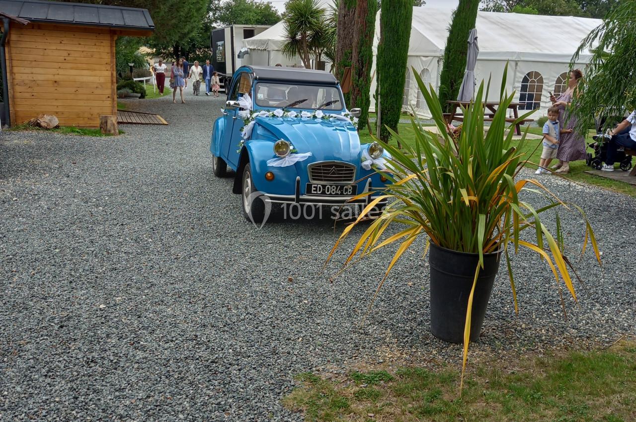 Une voiture ancienne bleue circule sur une allée gravillonnée bordée de végétation et de structures blanches.