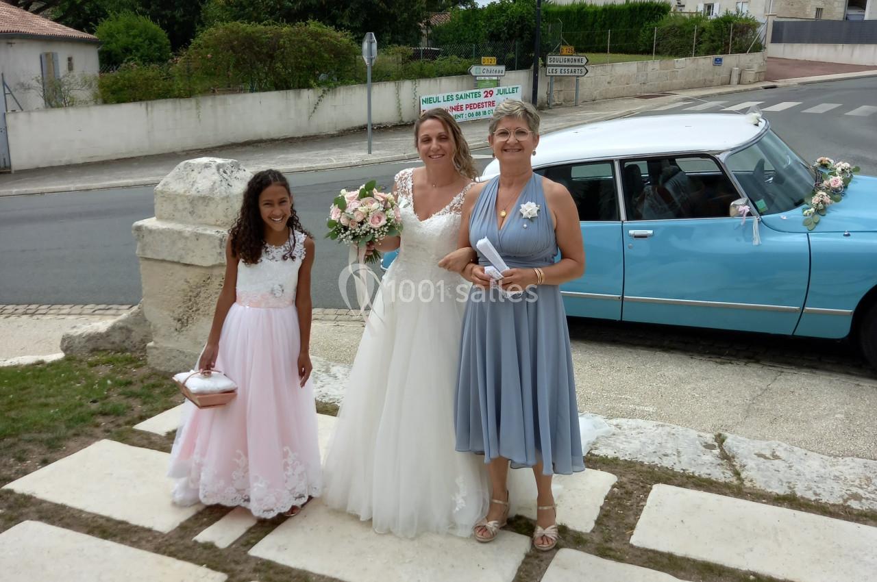 Une mariée souriante entourée de deux femmes tenant des bouquets, devant une voiture ancienne bleue dans un village.