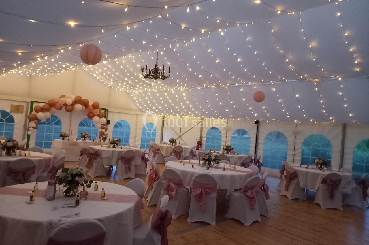 Salle de réception sous chapiteau décorée de guirlandes lumineuses, ballons et tables dressées avec nappes blanches et…