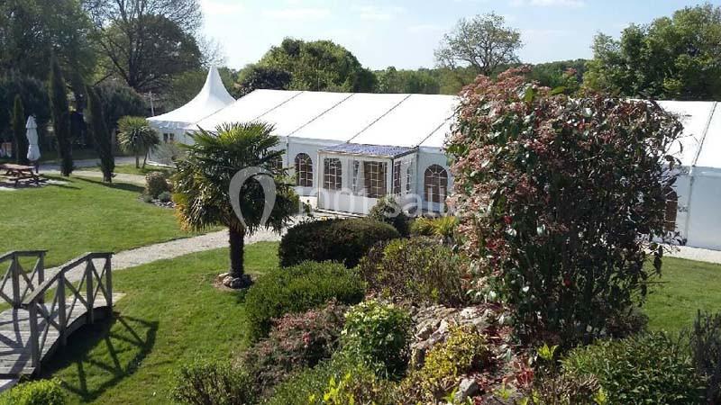 Vue d'un grand chapiteau blanc entouré de verdure, avec un jardin paysager et un pont en bois au premier plan.