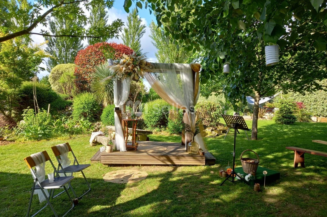 Arche de cérémonie décorée de voilages et de fleurs dans un jardin verdoyant avec chaises et accessoires disposés autour.