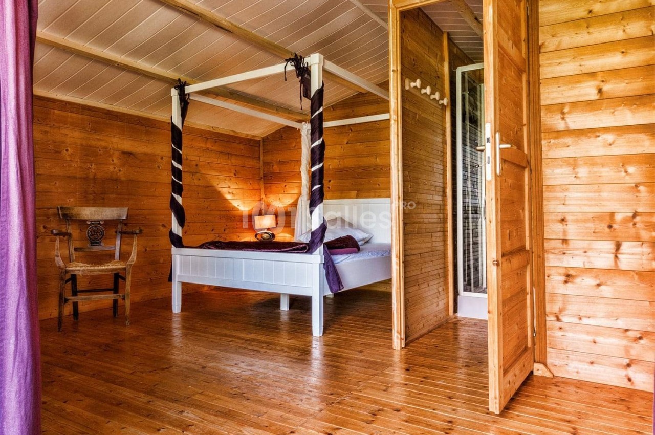 Chambre en bois avec lit à baldaquin blanc, chaise en bois sculpté et lumière naturelle entrant par une fenêtre.