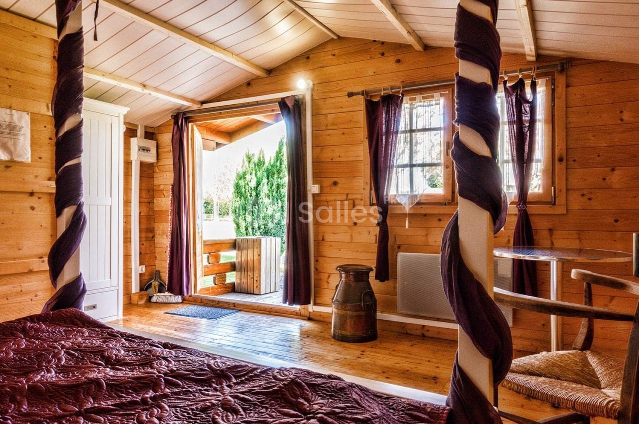 Chambre en bois avec lit à baldaquin, rideaux violets, table en bois et porte ouverte sur un jardin verdoyant.
