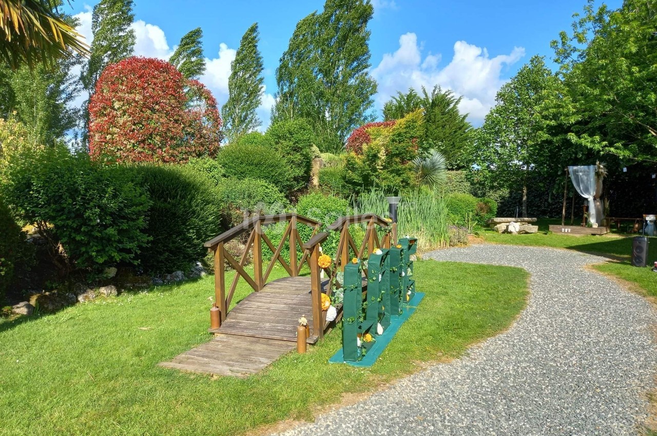 Petit pont en bois traversant une pelouse verdoyante, entouré d'arbustes et d'arbres sous un ciel bleu.