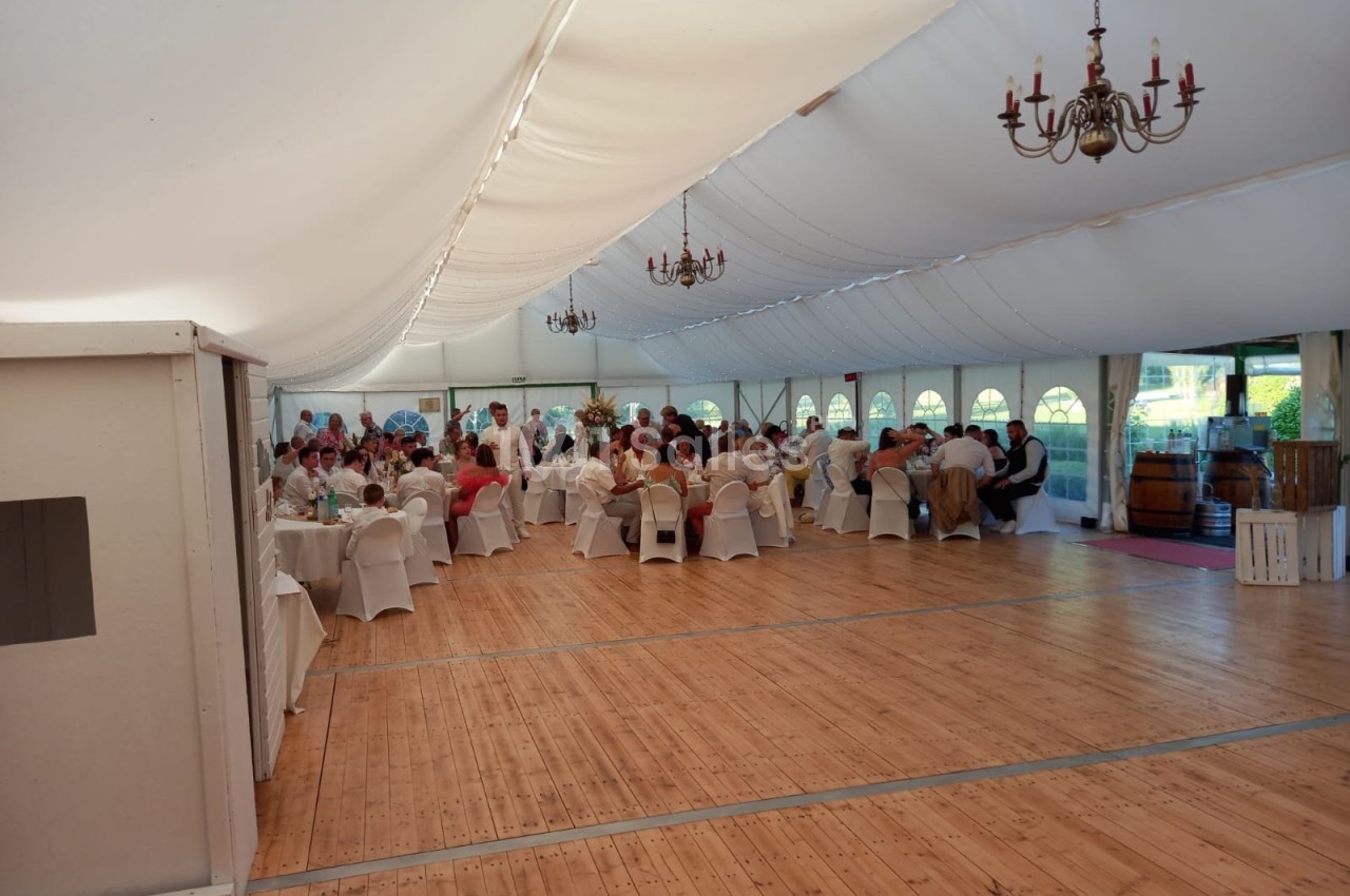Salle de réception sous une tente blanche, avec des invités assis à des tables rondes et un parquet en bois.