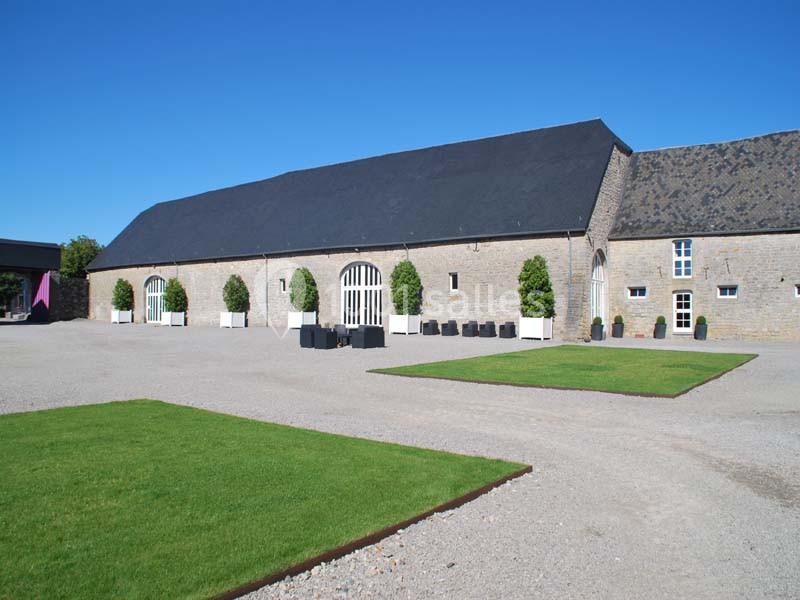 Location salle Ciney (Province de Namur) - Le Domaine d'Achêne #3 Cour extérieure avec pelouses rectangulaires, graviers et bâtiment en pierre à toit d'ardoise sous un ciel bleu.