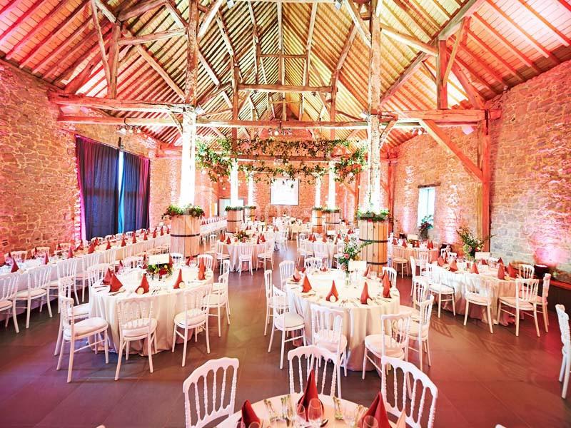 Location salle Ciney (Province de Namur) - Le Domaine d'Achêne #2 Salle de réception en pierre avec poutres apparentes, tables rondes dressées et décorations florales suspendues.