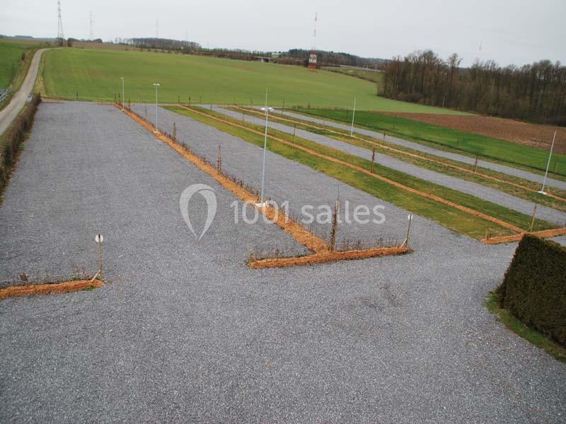 Location salle Ciney (Province de Namur) - Le Domaine d'Achêne #16 Parking en gravier vide, délimité par des haies et entouré de champs et d'arbres sous un ciel nuageux.