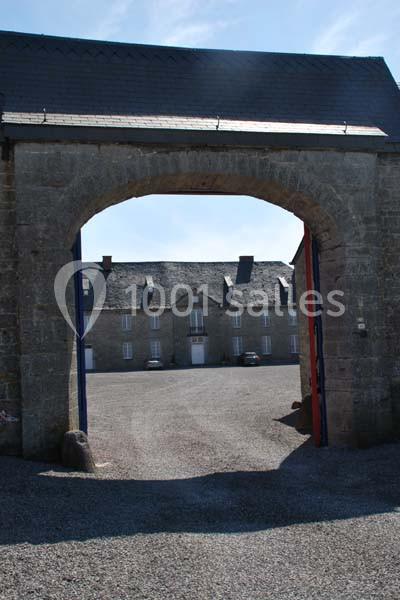 Location salle Ciney (Province de Namur) - Le Domaine d'Achêne #17 Vue d'une cour pavée à travers une arche en pierre, avec des bâtiments en arrière-plan sous un ciel dégagé.