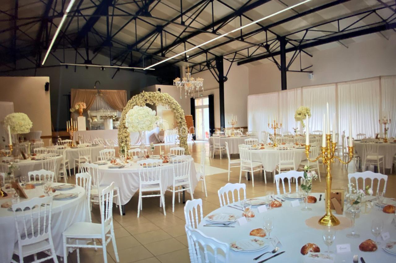 Salle de réception décorée pour un mariage avec tables rondes, nappes blanches, arche florale et chandeliers dorés.