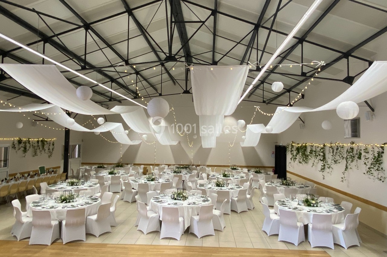 Salle de réception décorée avec des tables rondes, nappes blanches, guirlandes lumineuses et suspensions en tissu.