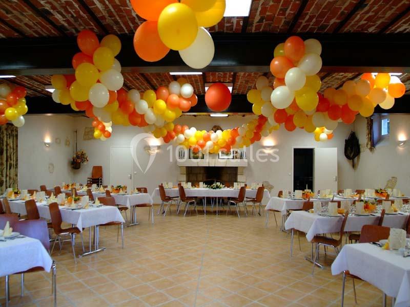 Salle décorée avec des ballons orange, jaunes et blancs, tables dressées pour un événement festif.