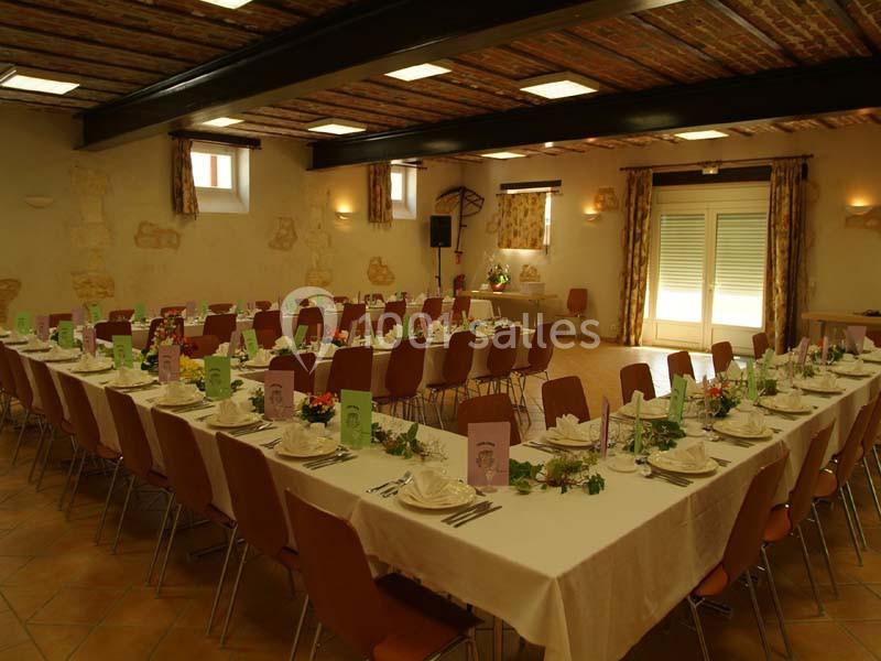 Salle de réception avec tables dressées en U, décorées de fleurs et menus colorés, éclairée par des fenêtres et des lampes.