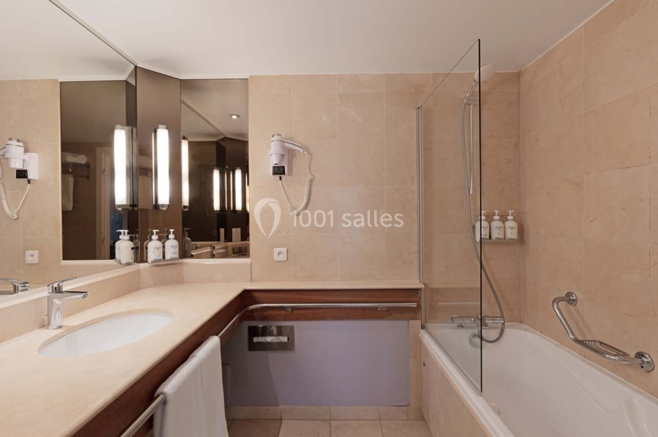 Salle de bain avec lavabo, miroir éclairé, baignoire avec paroi vitrée, sèche-cheveux mural et distributeurs de savon.