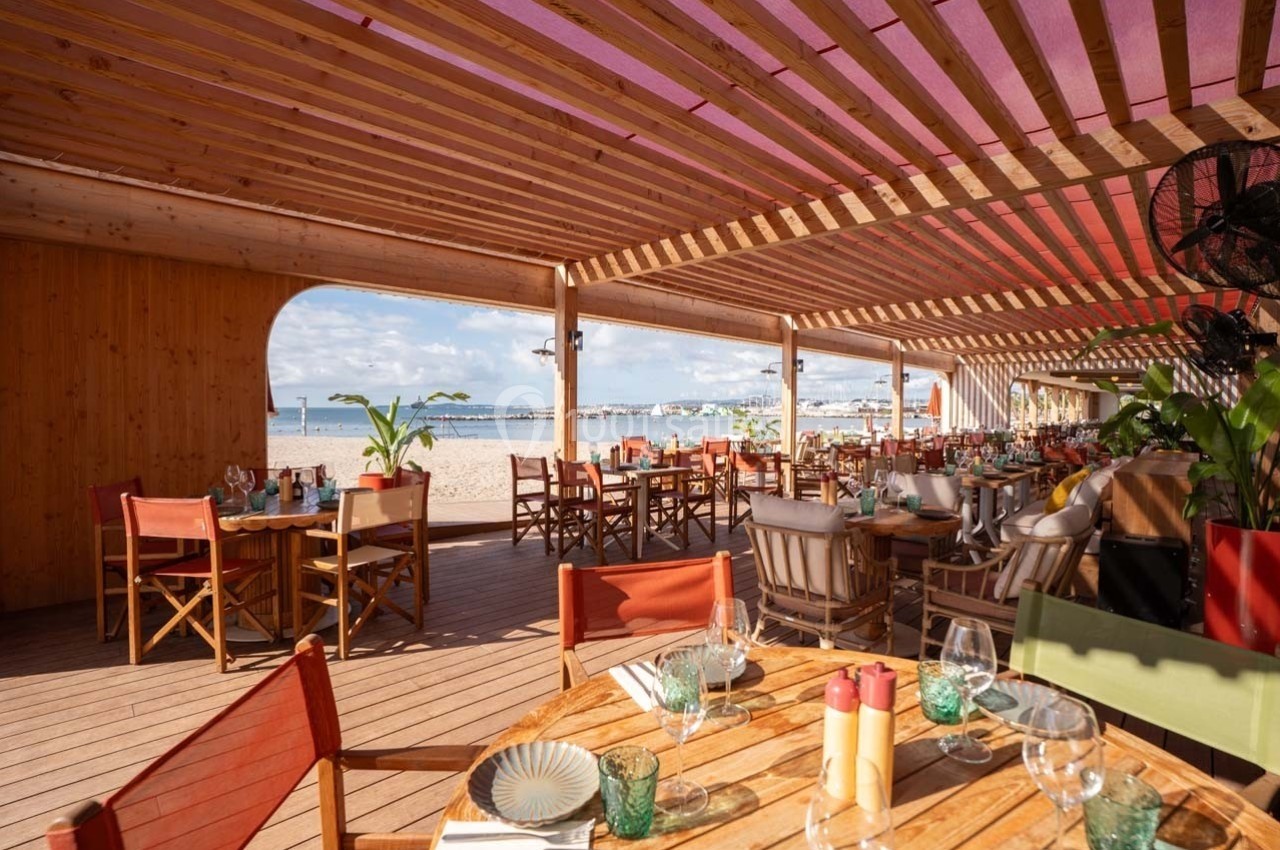 Terrasse d'un restaurant en bois avec vue sur la plage et la mer, tables dressées sous une pergola colorée.