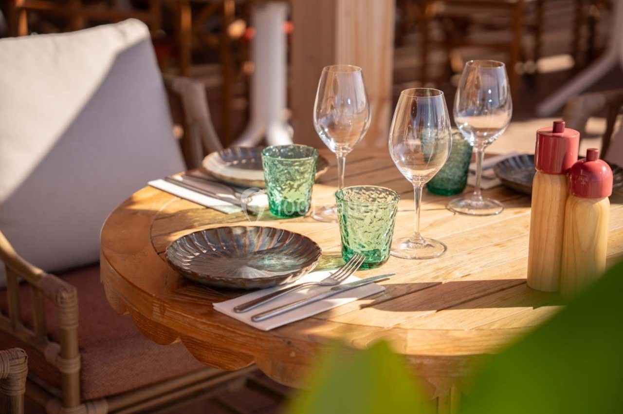 Table en bois dressée avec assiettes, verres, couverts et condiments, située en extérieur avec des chaises en rotin.