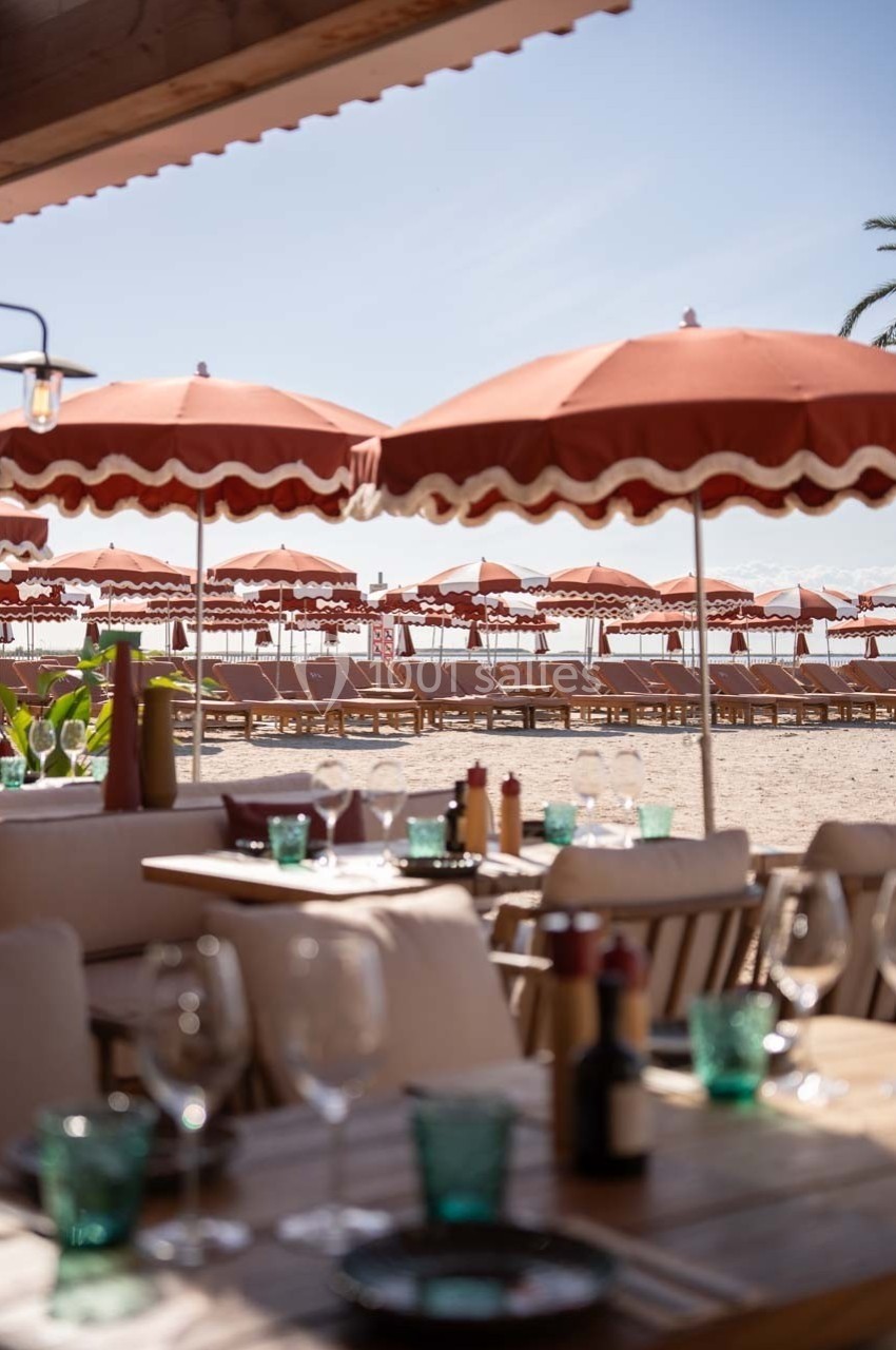 Terrasse d'un restaurant en bord de plage avec vue sur des parasols rouges et des transats alignés sur le sable.