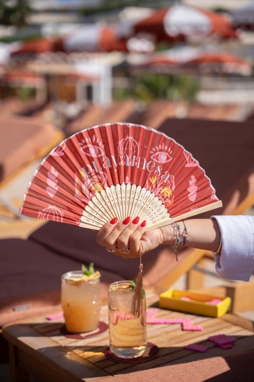 Main tenant un éventail rouge décoré, avec des cocktails et des transats en arrière-plan.