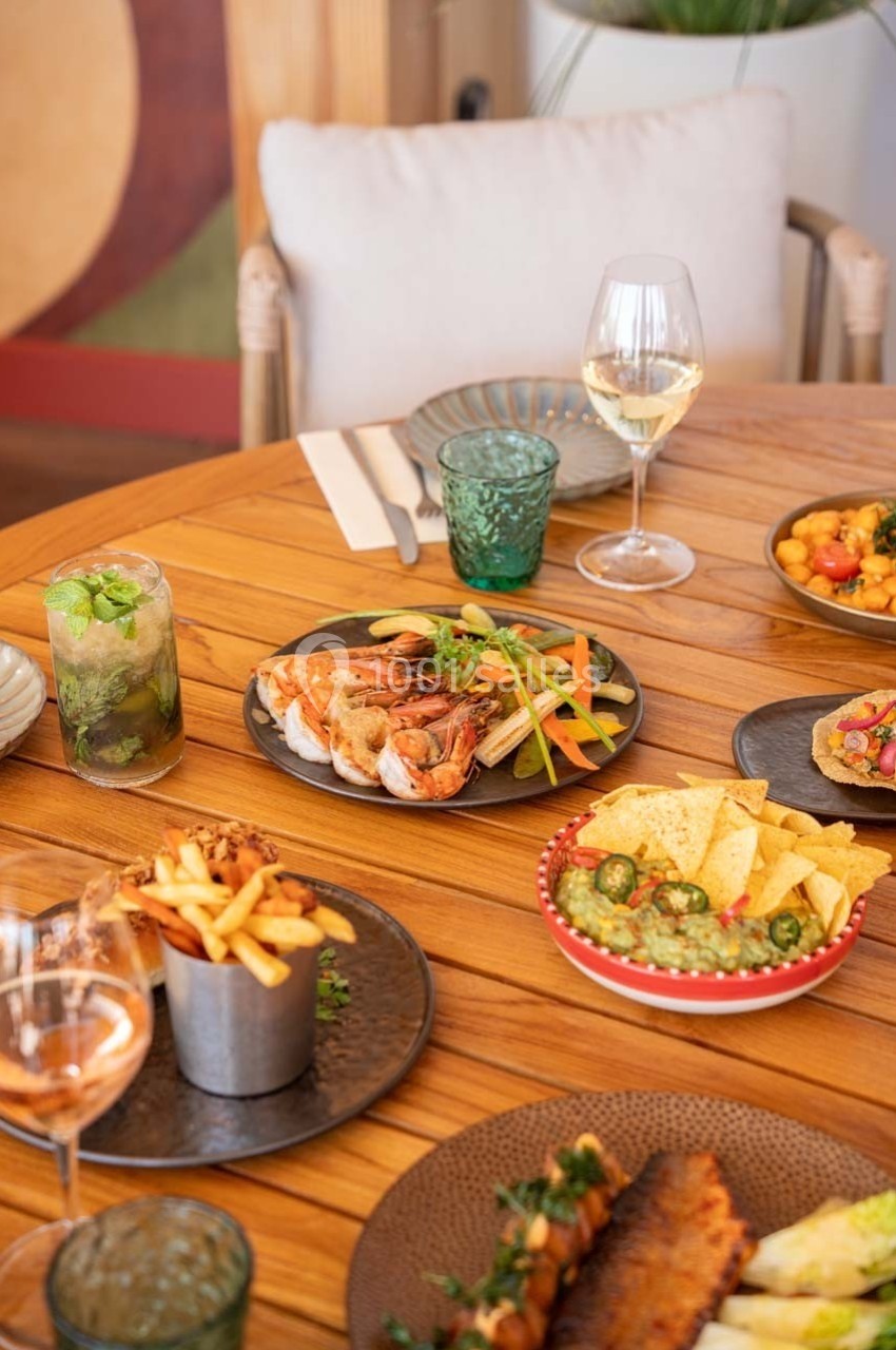 Table en bois avec des plats variés : fruits de mer, frites, nachos, guacamole, boissons et verres de vin.
