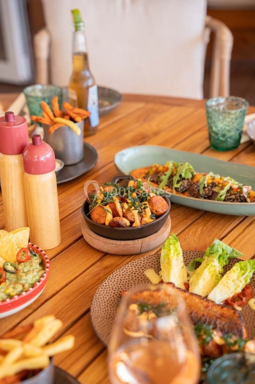 Table en bois garnie de plats variés, dont des légumes grillés, des salades, des frites et des sauces.