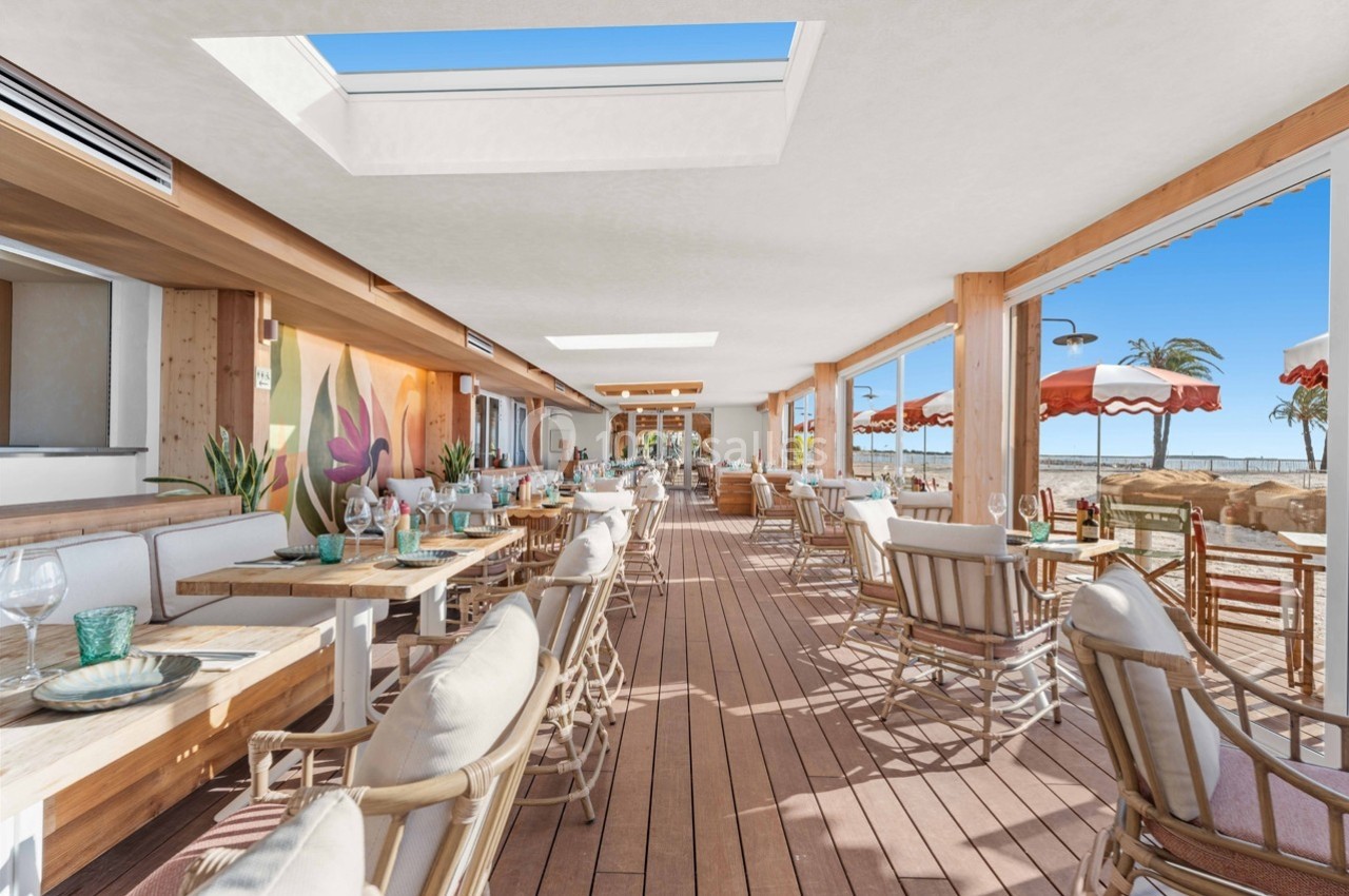 Salle de restaurant lumineuse avec terrasse en bois, tables dressées, grandes baies vitrées et vue sur la plage.