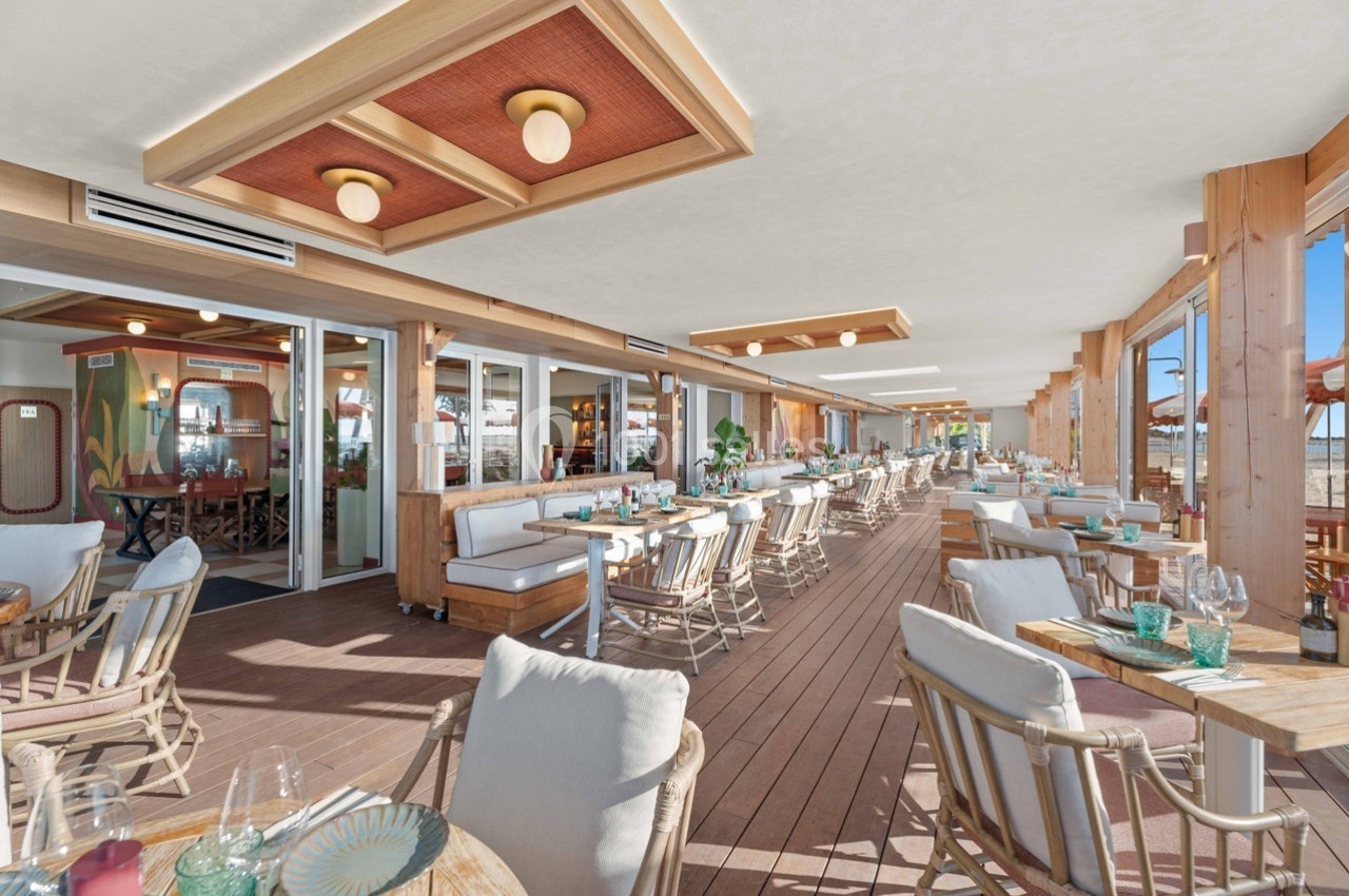 Terrasse couverte d'un restaurant en bord de mer avec mobilier en bois, tables dressées et vue sur la plage.
