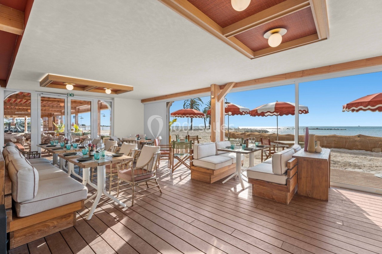 Salle de restaurant lumineuse avec terrasse en bois, vue sur la plage et parasols rouges en arrière-plan.