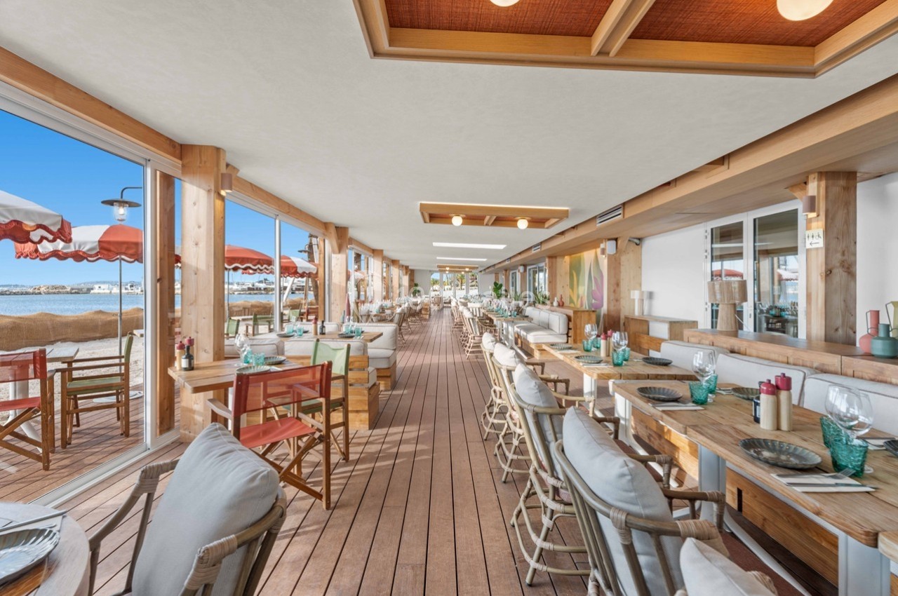 Salle de restaurant en bord de plage avec tables en bois, chaises confortables et vue sur la mer.